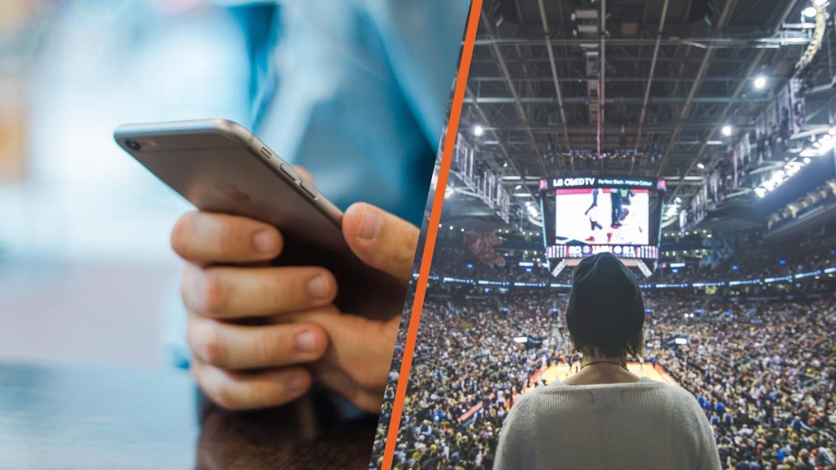 Una mano con un móvil junto a un aficionado que sigue un partido de baloncesto en un pabellón lleno.